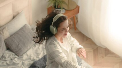 Energetic woman jumping on bed bedroom dancing hold mobile phone smartphone cellphone home headphones listen music technology active female girl enjoy sing song pastime weekend positive delight tenant