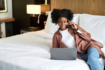Portrait of young African American woman using laptop and phone on bed in hotel room during...