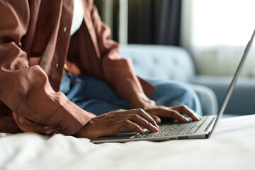 Side view closeup of African American young woman typing on laptop keyboard while sitting on bed in...