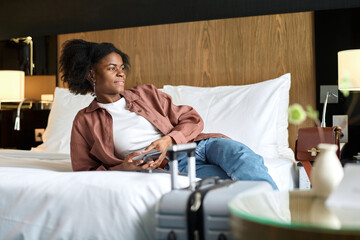 Portrait of young African American woman relaxing on bed in hotel room upon arrival and looking at...