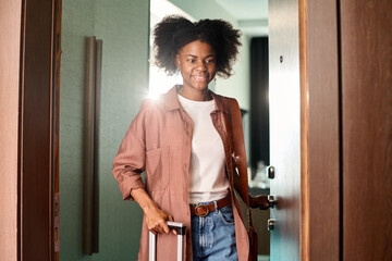 Waist up portrait of smiling African American woman entering hotel room with suitcase copy space