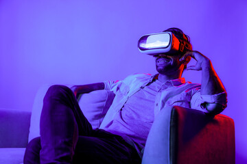 Happy man sitting at sofa while wearing VR goggle to watch funny movie. Caucasian person relaxed...