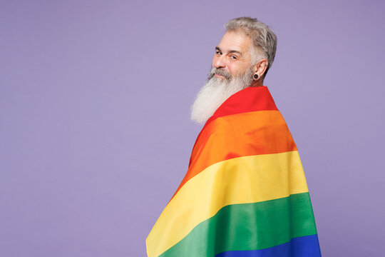 Side view young smiling elderly bearded gay man 50s years old wears casual clothes wrapped in striped rainbow flag look camera isolated on plain purple background. Lifestyle LGBT June pride concept. - Powered by Adobe