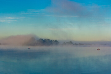 Fishermen on  boats early in the morning at dawn in the fog at golden hour catch fish on the Oka River, Russia. Barge, tugboat, green grass and trees, a river bend against a clear light sky in summer.