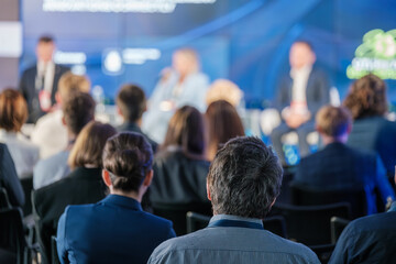 Attendees watch and listen to a panel of speakers discussing topics at a professional business conference. The focus is on the engaged audience.