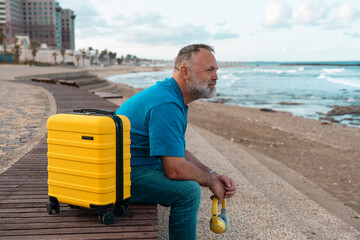 bearded mature man with suitcase and headphones sitting on bench looking at sea on summer day