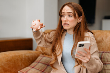 redhead woman with grimace of displeasure holding piggy bank and phone saving money for rainy days...