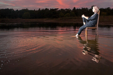 A person peacefully enjoys a beautiful sunset while sitting on a chair partially submerged in calm...