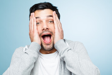 Man, shocked or surprised in portrait with hands on face in blue studio background for excitement,...