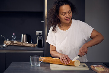 Mixed race beautiful woman in white t-shirt, standing at kitchen counter, holding knife and slicing...