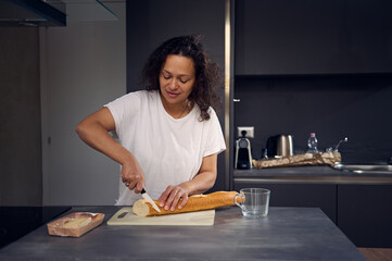 Multi ethnic pretty woman in white t-shirt, standing at kitchen counter, holding knife and slicing...