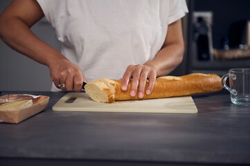 Close-up hands of a woman in white t-shirt, standing at kitchen counter, holding a kitchen knife and cutting a loaf of bread on cutting board, preparing breakfast in the home kitchen in the morning