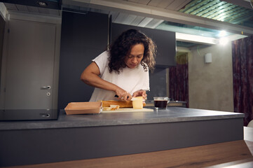 Curly haired mixed race adult woman slicing cheese on cutting board, using a kitchen knife,...