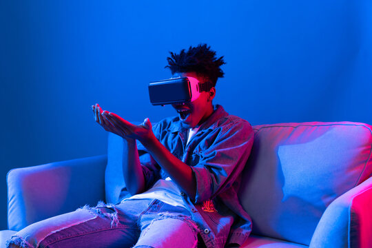 Young African American looking VR enjoy watching fantastic 3D pointing interesting object technology blue neon light virtual reality metaverse new world at living room sitting on sofa. Contrivance.