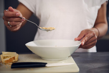 Details on the hand of woman in white t-shirt, holding a spoon with wholegrain muesli granola above a white bowl, enjoying a healthy breakfast in the morning