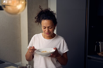 Young pretty woman in white t-shirt, holding a bowl of muesli, standing at kitchen counter at home....