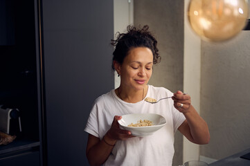 Young woman in white t-shirt, enjoying her wholesome breakfast, eating a bowl of muesli, standing...