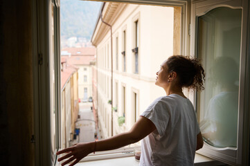 Rear view. Young woman in white t-shirt, opening windows in the living room, enjoying the morning...