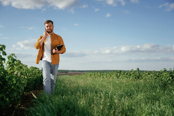 Talking by phone, holding notepad. Handsome man is on the agricultural field at daytime