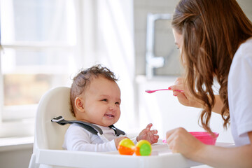 Mother, kitchen and feeding baby with high chair for meal, snack time or breakfast at home. Happy...