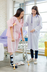 Asian female patient with broken leg assisted by woman doctor. Injured young woman stands up from...