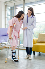 Asian female patient with broken leg assisted by woman doctor. Injured young woman stands up from...