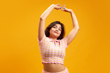 Charming, young brunette Indian woman raising hands while dancing against vibrant yellow studio...