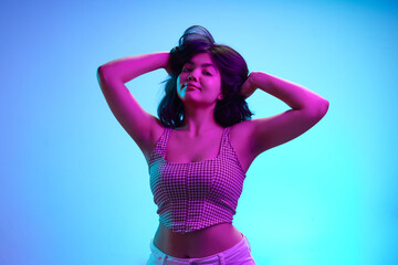 Portrait of beautiful Indian woman posing raising hands in pink neon light against gradient blue background. Concept of human emotions, self expression, beauty and fashion, youth. Ad