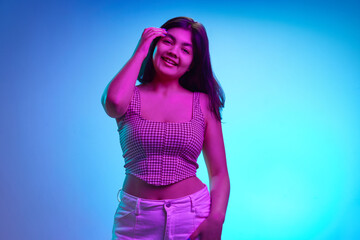 Portrait of young Indian woman looking at camera and cheerful smiling in pink neon light against gradient blue background. Concept of human emotions, beauty and fashion, youth. Ad