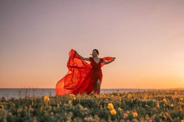 woman red dress is standing on a grassy hill overlooking the ocean. The sky is a beautiful mix of...