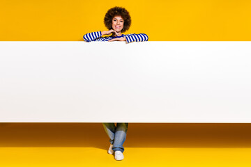 Full length photo of cheerful woman dressed striped shirt standing behind white placard empty space...