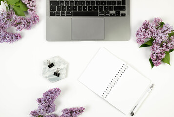 Top view of purple lilac flowers on white background. Laptop computer, white notebook, pen, binder...