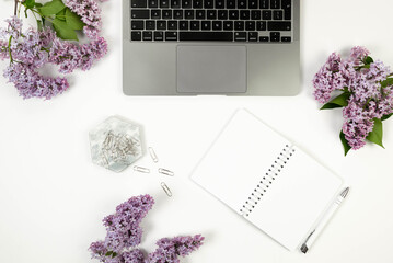 Top view of purple lilac flowers on white background. Laptop computer, white notebook, pen, paper...