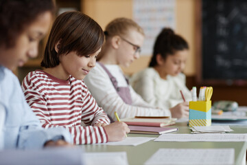 Selective focus shot of Caucasian boy wearing casual clothes sitting at table with his classmates doing writing exercise