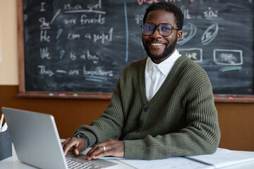 Medium portrait of young African American male teacher of English wearing eyeglasses sitting at...