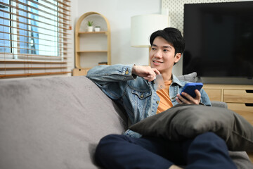 Attractive asian man relaxing on couch at home holding mobile phone and looking outside window