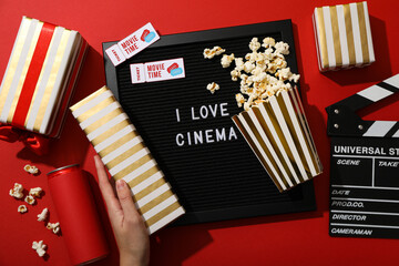 Black letter board with text, clapperboard, popcorn and hand with gift boxes on red background, top...
