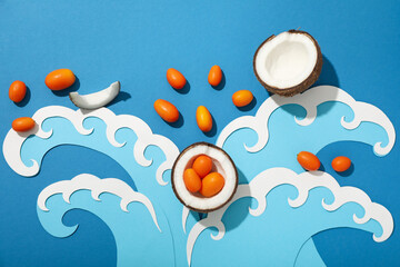 Pieces of coconut, whole kumquats and paper waves on blue background, top view