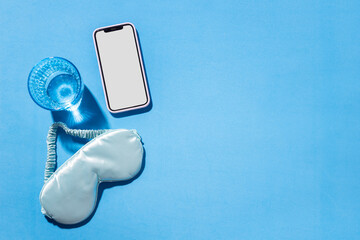 Glass of water, sleeping mask and smartphone on blank blue background. Concept of good sleep and tracking of sleep