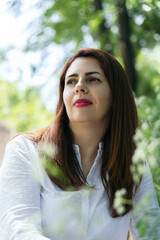 Portrait of a 45 years old mature woman with red lips, looking to the side happy and confident in an outdoor park on a sunny day. Concept of maturity, happiness and well-being