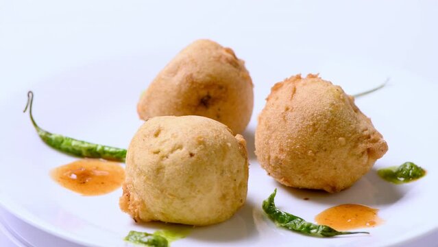 Potato fritters, potato dumplings (fried potatoes in round shape) Batata vada or bonda, on a white plate with green chillies and red tomato sauce . Plate rotates with the cooked potatoes. Indian food