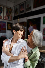 Two mature women wrap each other in a warm hug in an artistic studio.