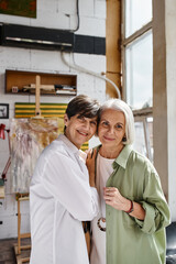 A mature lesbian couple standing side by side in an art studio.