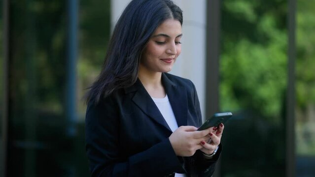 Successful young indian businesswoman hold smartphone scrolling social media texting browsing online checking email during break at work and looking ahead in modern business centre