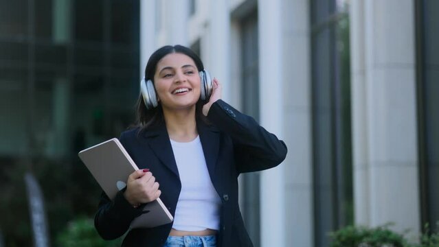 Charming young indian businesswoman with headphones listening to music while walking down the street after work outdoors Happy relaxed lady on the city centre enjoying beautiful day alone