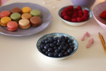 Plate of pastel macarons, cookies and chocolate, cup of tea of coffee, glass of bubble water, various berries, books and accessories on the table. Selective focus, pastel colors.