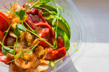 Grilled shrimps, red bell pepper with avocado and tomato in plastic bowl. Poke bowl in sunlight....