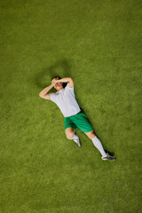 Aerial view of soccer player in green shorts and white jersey lying on grass, covering his face...