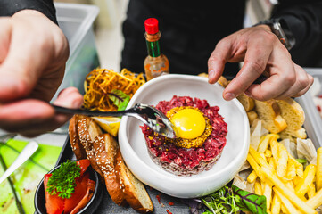 Close-up of a hand holding a fork over a plate of steak tartare topped with a raw egg yolk, surrounded by fries, bread, and greens
