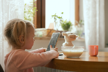 Baby girl eats breakfast and looks at the phone at the table. Harmful upbringing of a child with...